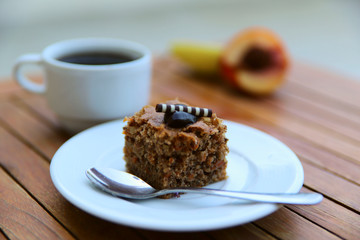 Blurred food background. On a wooden table is a white plate with a piece of chocolate cake and a spoon, in the background is a cup of coffee and fruits. Close-up, side view, horizontal. Food concept.