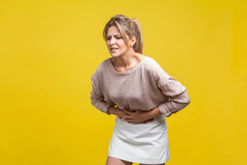 Portrait of unhealthy young woman with fair hair in casual beige blouse standing, touching stomach...