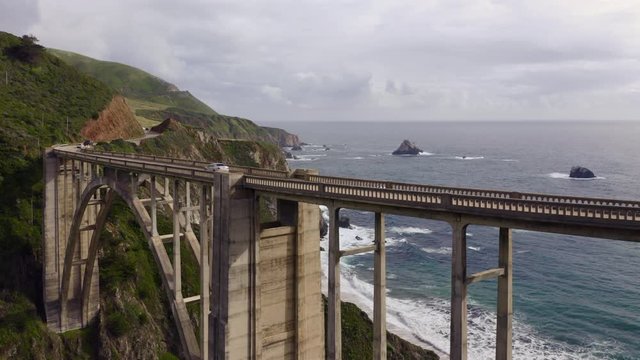Impressive aerial footage of the unique construction of Bixby Bridge. Drone moves slowly towards the beautiful arch bridge, crosses it above and overlooks the stunning coastline of Pacific ocean. 4K