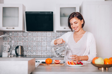 Young sincerely smiling female pouring a purified mineral water into transparent glass decanter at...