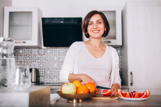 Beautiful young sincerely smiling female chopping grapefruit using a knife and cutting board in modern kitchen. Plenty of apples, grapefruits, kaki and oranges fruits, berries are on the tables.