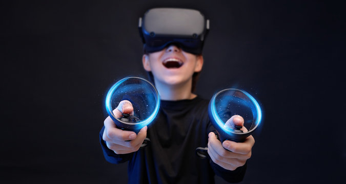 Young boy on dark background. Youngster using VR helmet with controllers in hands playing in video games and expresses happiness. Blue neon light.