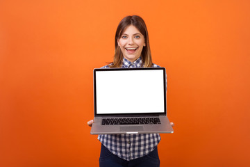 Naklejka premium Portrait of glad beautiful woman with brown hair in checkered shirt standing holding laptop with blank screen and smiling at camera, place for ad. indoor studio shot isolated on orange background