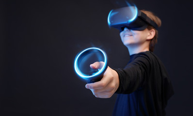 Young boy on dark background. Youngster using VR helmet with controllers in hands playing in video games and expresses happiness. Blue neon light.