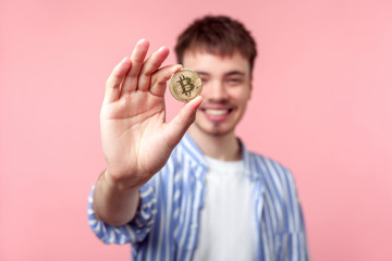Fototapeta premium Portrait of positive excited brown-haired man in casual shirt standing showing golden bitcoin and looking at camera with toothy smile, cryptocurrency. indoor studio shot isolated on pink background