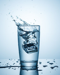 One glass of water with splashes from the fall of an ice cube on a light blue background