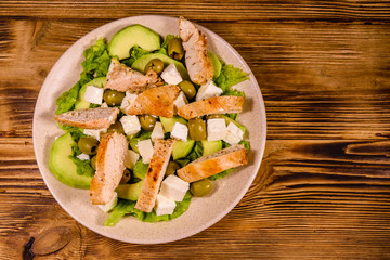 Fresh salad with chicken meat, feta cheese, avocado, green olives and lettuce leaves in ceramic plate on wooden table. Top view