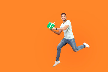 Full length portrait of excited joyous brunette man in sneakers and denim outfit smiling at camera while flying or running in air with gift box, present. studio shot isolated on orange background