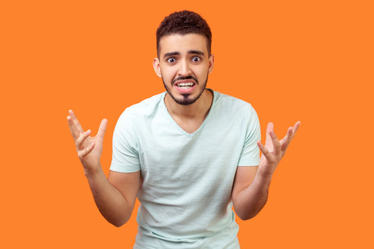 How Could You? Portrait Of Annoyed Frustrated Brunette Man With Beard In White T-shirt Standing With Raised Hands And Indignant Face Asking Why. Indoor Studio Shot Isolated On Orange Background