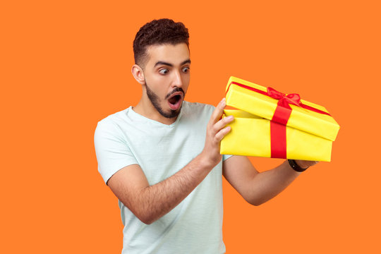 What's in the box. Portrait of curious man with beard in white t-shirt looking inside gift box, peeking with interest and amazement, unpacking long awaited present. studio shot, orange background