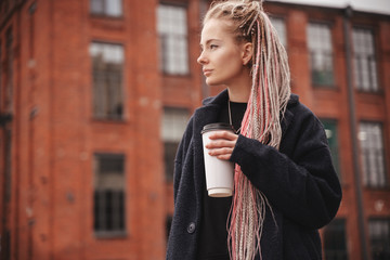 Stylish beautiful young blonde girl with pink dreadlocks with a white glass of take-away coffee,...