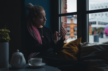 Stylish beautiful young blonde girl with pink dreadlocks sits in a cozy cafe with a smartphone in her hands and drinks tea