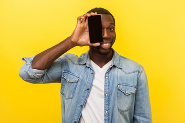 Portrait of happy cheerful man in denim casual shirt covering half face with cellphone and looking at camera with toothy smile, positive glad face. indoor studio shot isolated on yellow background