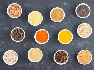 Buckwheat, millet, allspice, fennel, oat, pepper chili, masala, fenugreek, cloves, turmeric, cumin, mustard seeds, green cardamom in white bowls on black concrete background.