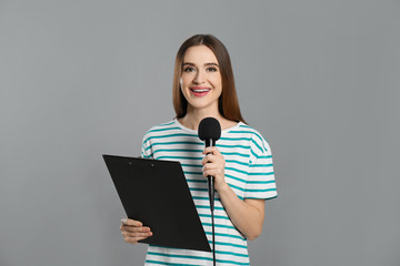 Young female journalist with microphone and clipboard on grey background