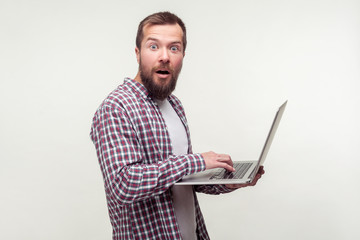 Portrait of amazed computer user, bearded man in casual plaid shirt standing with laptop and looking at camera with shocked expression, excited surprised about email news. studio shot white background
