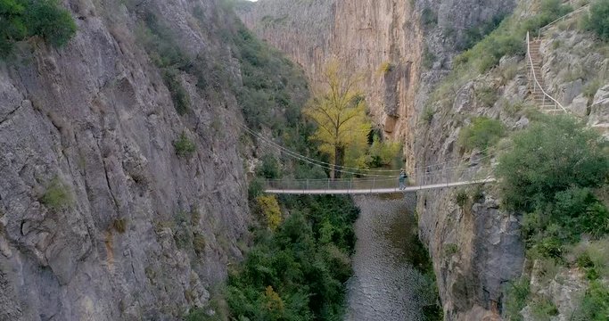 Vuelo descendiendo de dron dentro de desfiladero de roca y al fondo jubilado senderista cruzando puente colgante