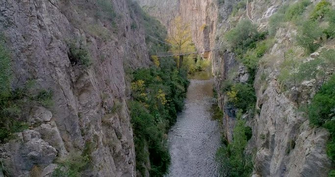 Dron avanzando por un ca&ntilde;&oacute;n de rio profundo y al fondo un puente colgante y un anciano lo cruza