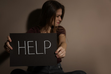 Abused young woman with sign HELP near beige wall. Domestic violence concept