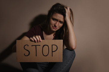 Crying young woman with sign STOP near beige wall. Domestic violence concept