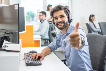 Smiling call center operator in headset looking at camera and showing thumb up