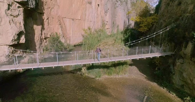 Vuelo hacia atr&aacute;s de dron en garganta de r&iacute;o profunda y persona mayor caminando en puente colgante