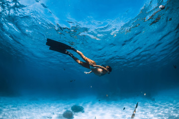 Freediver girl with fins glides over sandy bottom with fishes in blue ocean
