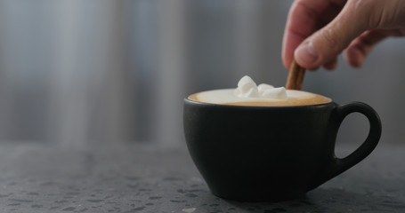 adding cinnamon stick into cappuccino in black cup on terrazzo countertop
