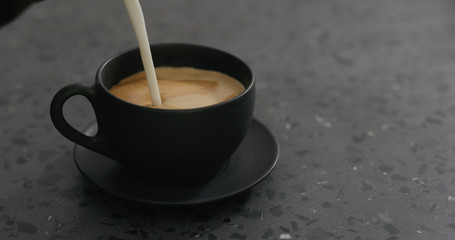 pouring steamed milk into cappuccino in black cup on terrazzo countertop