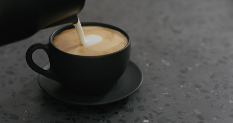 pouring steamed milk into cappuccino in black cup on terrazzo countertop