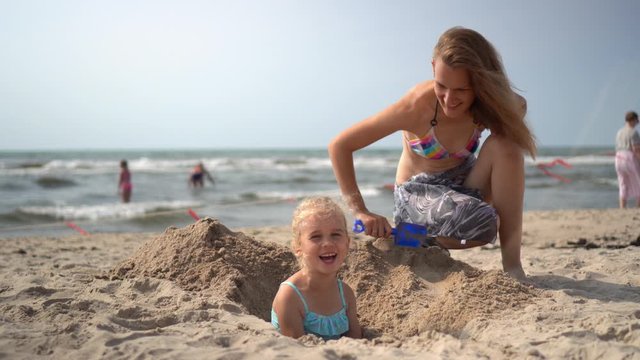 mother in bikini bury her little blond girl in the beach sand. camera movement