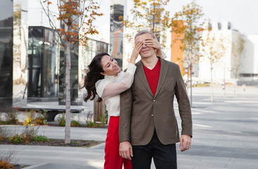 Beautiful Asian woman prepared surprise for her male friend and closed his eyes on the street. Fashionable couple wearing stylish business clothes (Red pants, brown jacket, Burgundy jumper)