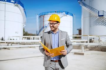 Handsome caucasian businessman in suit standing outdoors and holding folder. In background are oil storage tanks.