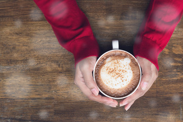 Woman's hands in red cadigan holding cup of cappucino coffee in winter.