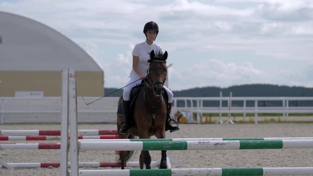 Horsegirl riding strong brown horse jumping over obstacles in outdoors sandy parkour riding arena. Female equestrian rider running on stallion at show jumping competition. Close up, slow motion.