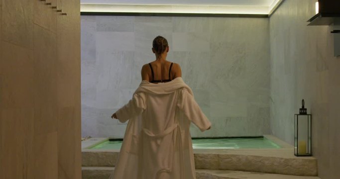 Back view of an young woman with firm and slim body is taking off a white bathrobe and entering in a whirlpool bath tube in a luxury wellness center.
