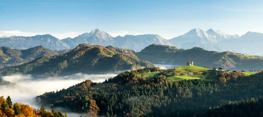 Sveti Tomaz church with low lying fog with the alps in the background during sunrise in fall season Slovenia