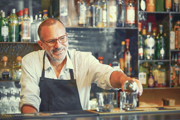 Portrait of professional elderly bartender man pours whiskey to the client of the hotel bar. The...