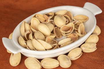 Lot of whole salted pistachio in small white ceramic bowl on cognac leather