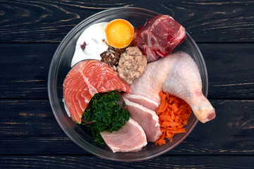 Different natural dog food in bowl on black wooden background. Meat fish egg vegetables bones organic food supplements. 