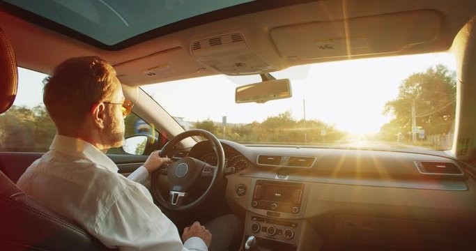 Back view of classic casual adult businessman in white shirt and sunglasses driving car with amazing sunlight rising in front of the road. Freelance traveling. Urban lifestyle. Navigation. Map. Driver