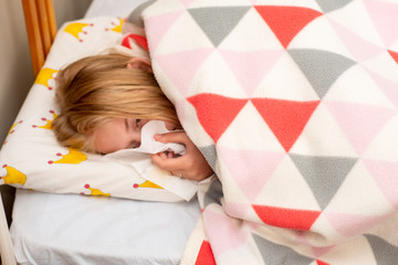 Obraz na płótnie Canvas Sick teenage girl laying in bed covered with blanket sneezing and blowing her nose with a tissue. Flu or allergy symptoms