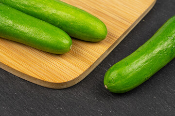 Group of three whole fresh green baby cucumber on bamboo cutting board on grey stone