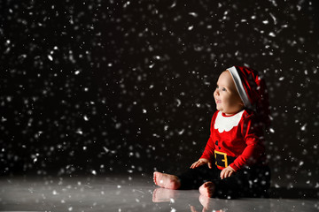 Smiling infant baby boy toddler in red christmas cap and new year costume is sitting on ice under the snow looking up