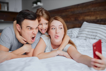 Obraz na płótnie Canvas young caucasian parents lying on bed with little daughter kid, use red smartphone and take selfie, emotional happy family