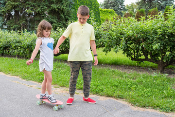 Big brother teaches little sister skateboarding in the park, holding her hand.