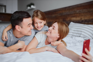 young caucasian parents lying on bed with little daughter kid, use red smartphone and take selfie, emotional happy family