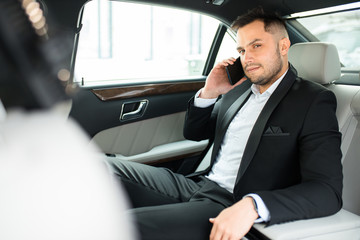 busy elegant male sitting inside of car and talking on phone, caucasian man with beard wearing tux
