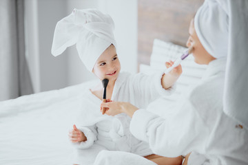 caucasian woman and little daughter sit on bed wearing towels and bathrobe doing makeup after shower. family concept