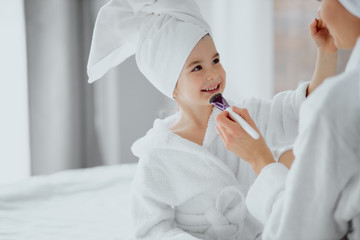 smiling happy positive cute little girl look at her mother and apply makeup with brushes, wearing white bathrobe and towel